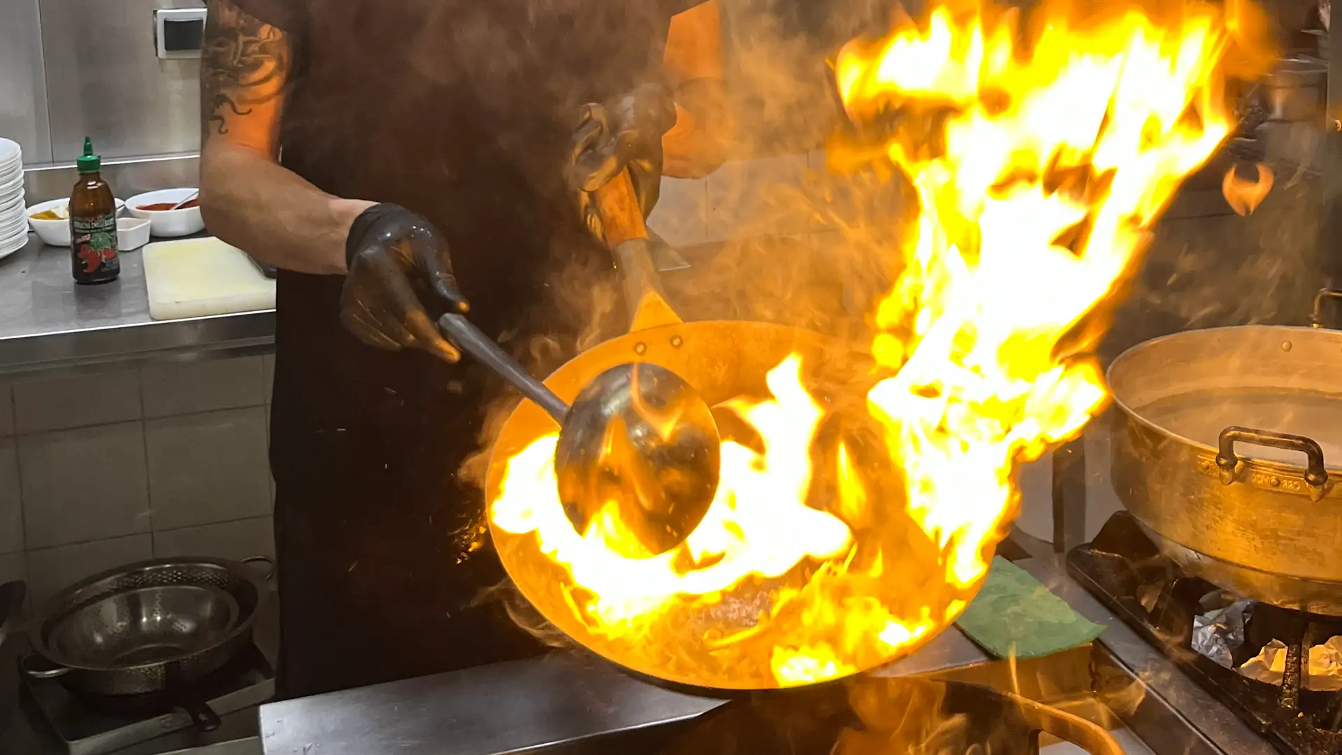 Un cocinero flamea el wok: las llamas suben por encima del borde mientras remueve con el cucharón